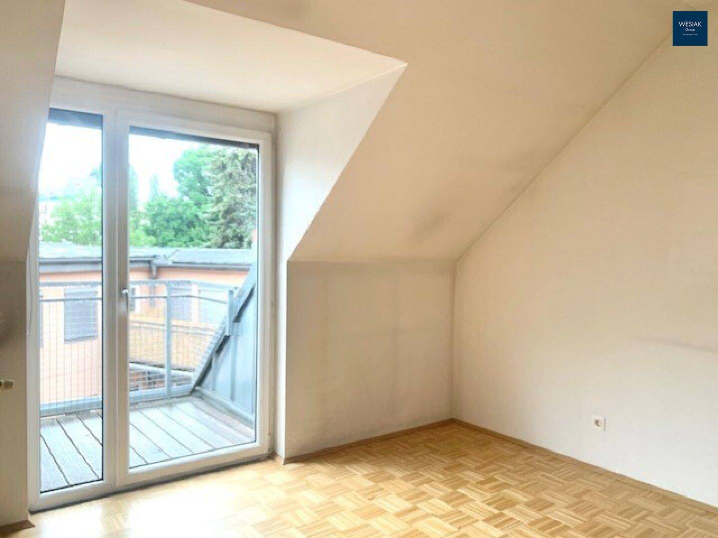Helles Zimmer mit Dachschräge, Parkettboden und Zugang zu einem Balkon.