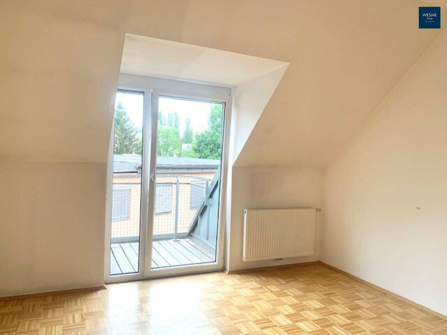 Geräumiges Zimmer mit Dachschräge, Parkettboden, Heizkörper und großem Fenster zum Balkon.