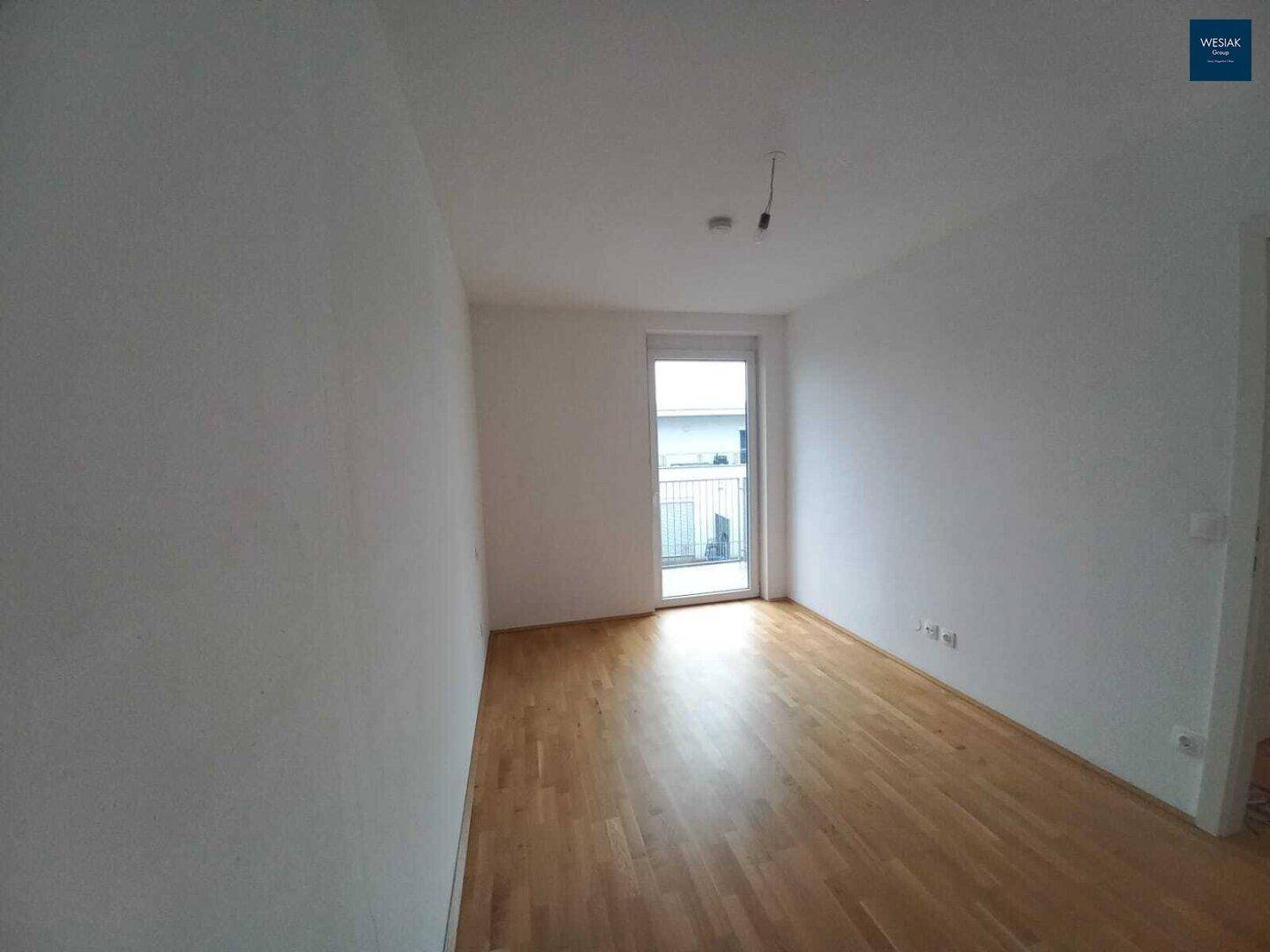 Leerer Raum mit Parkettboden und Zugang zu einem Balkon, hell und geräumig.