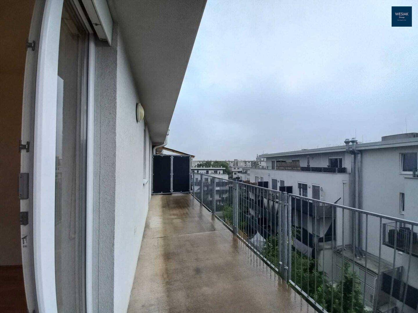 Langer Balkon mit Metallgeländer und Blick auf umliegende Gebäude, bei bewölktem Wetter.