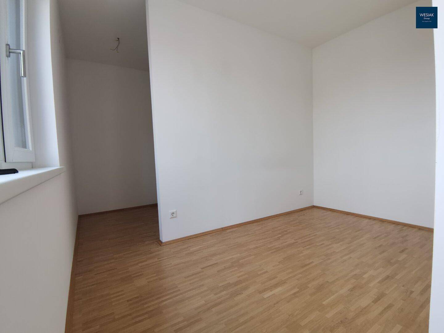 Leerer Raum mit Parkettboden und einem Fenster, das eine helle Atmosphäre schafft.