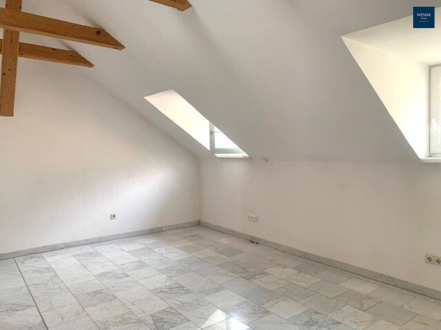 Helles Dachzimmer mit schrägen Wänden, Holzbalken und einem großen Dachfenster.