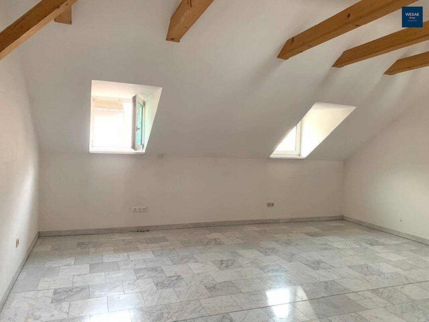 Leeres Dachzimmer mit schrägen Wänden, Holzbalken und zwei Dachgauben für natürliches Licht.