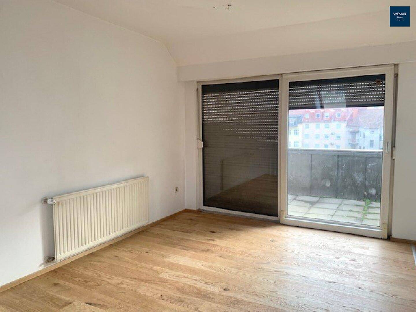 Leeres Zimmer mit Holzfußboden, Heizkörper und Zugang zu einem kleinen Balkon.