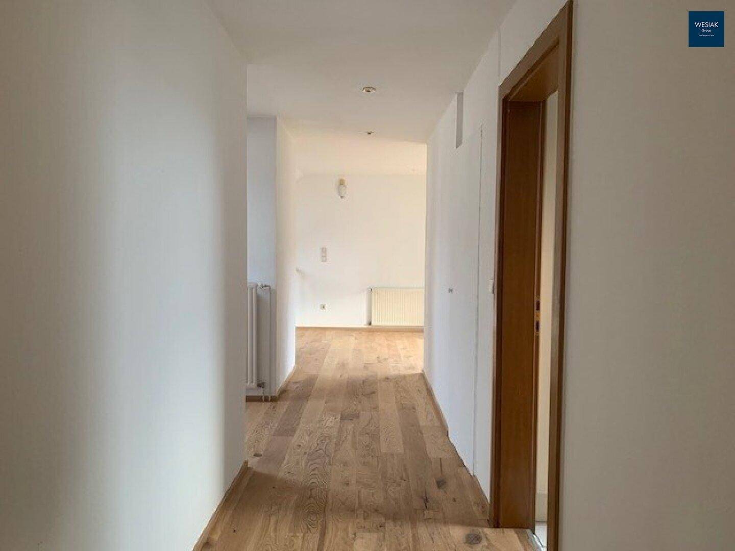 Langer, heller Flur mit Holzfußboden und weißen Wänden, der zu weiteren Räumen führt.