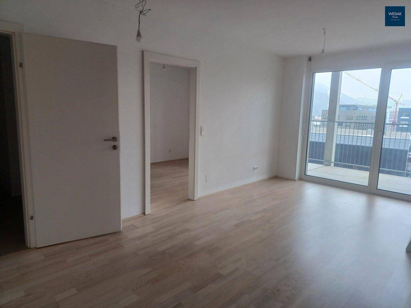 Heller, leerer Wohnraum mit hellem Parkettboden und Zugang zu einem Balkon.