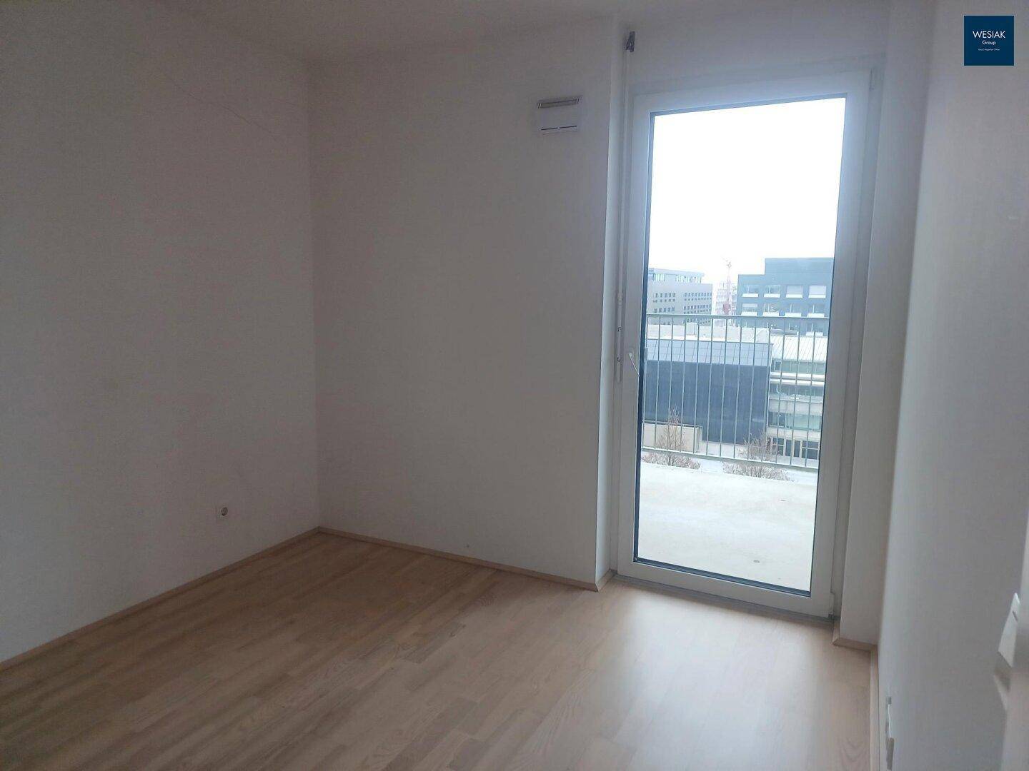 Leeres Zimmer mit hellem Holzboden und einer großen Fenstertür, die viel Licht hereinlässt.