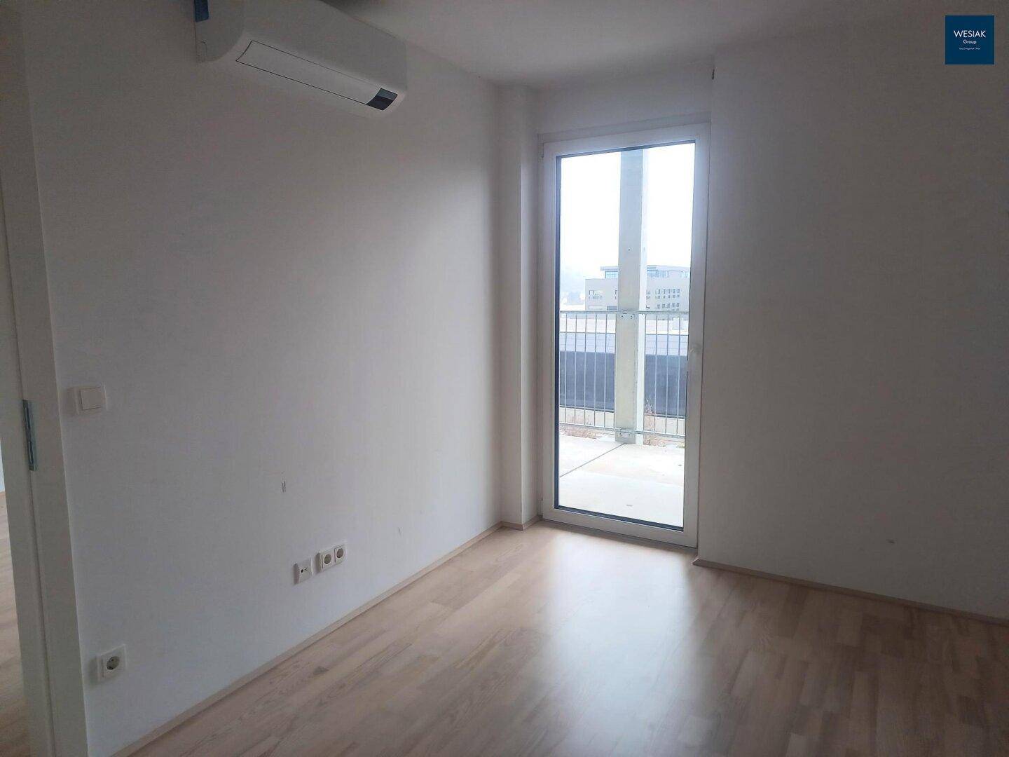 Helles, leeres Zimmer mit Holzboden und Zugang zu einem Außenbereich durch eine Fenstertür.