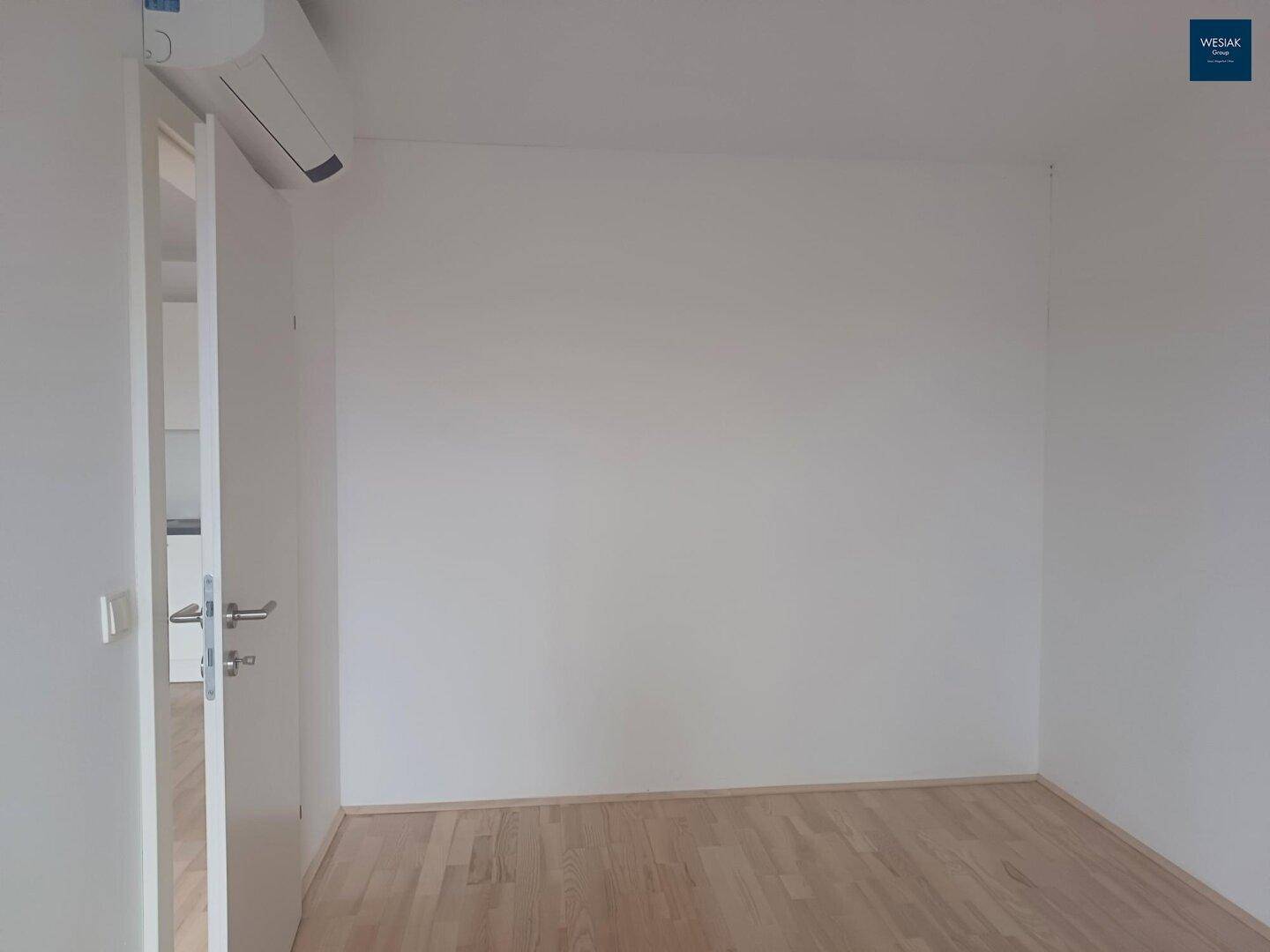 Leerer Raum mit weiß gestrichenen Wänden, hellem Holzboden und einer Klimaanlage an der Decke.