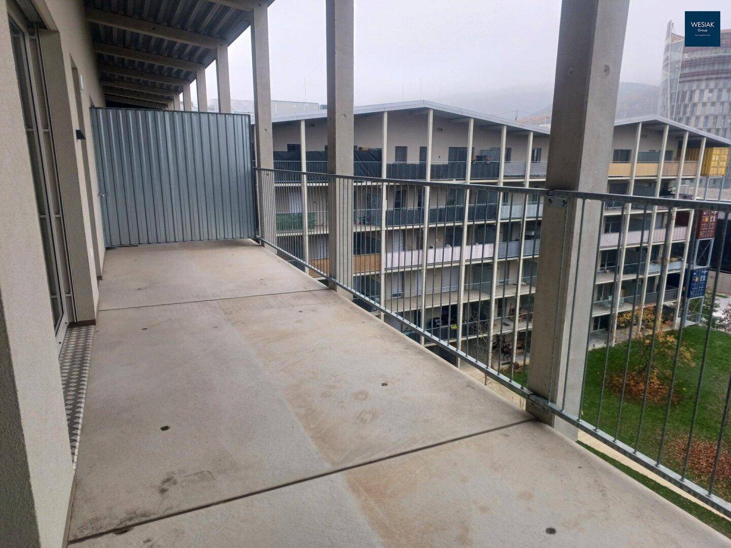 Geräumiger Balkon mit Betonboden und Metallgeländer, mit Blick auf andere Gebäudeteile.