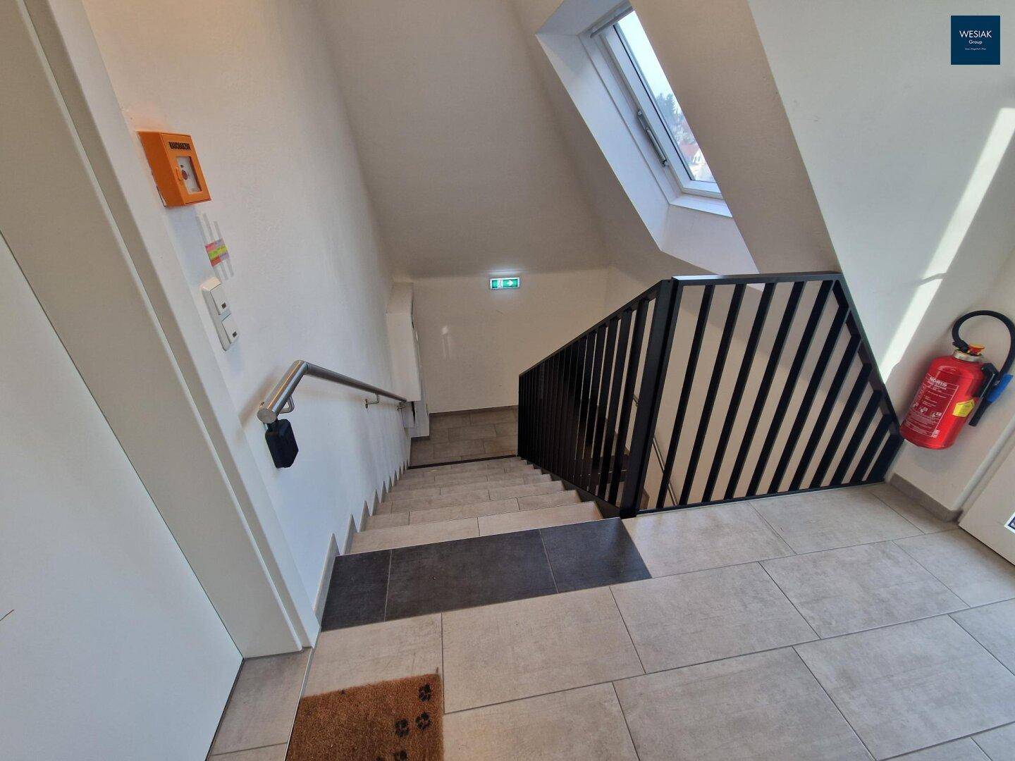 Heller Treppenbereich mit modernem Metallgeländer, Dachfenster und Fliesenboden, der nach unten führt.