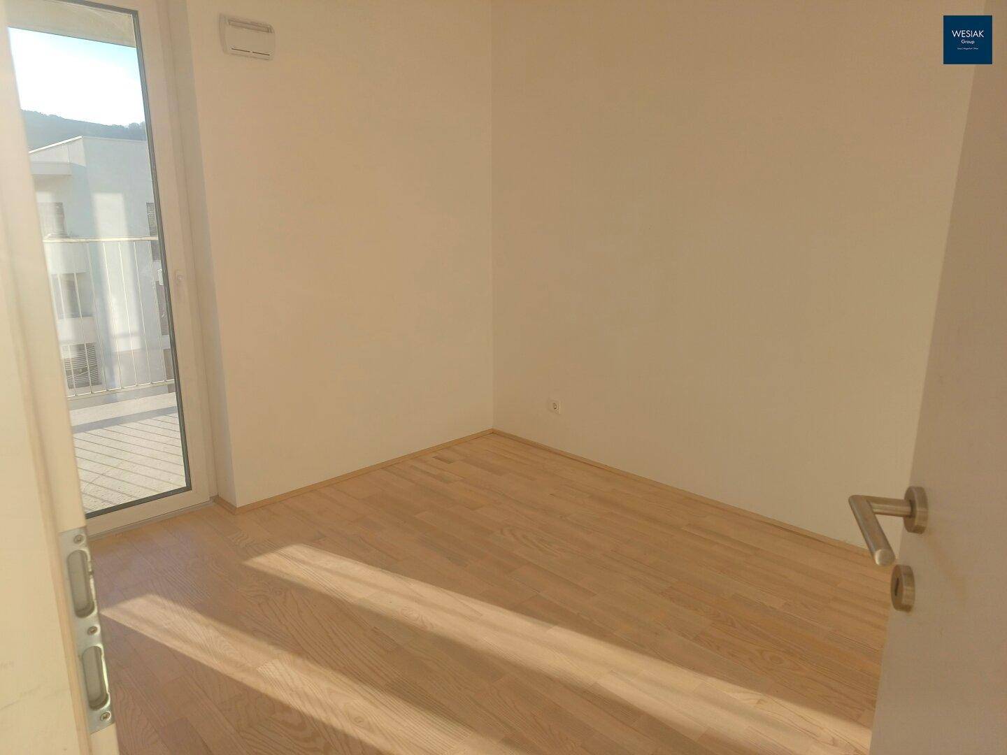 Helles Zimmer mit Holzboden und Zugang zu einem Außenbereich, durchflutet von Sonnenlicht.