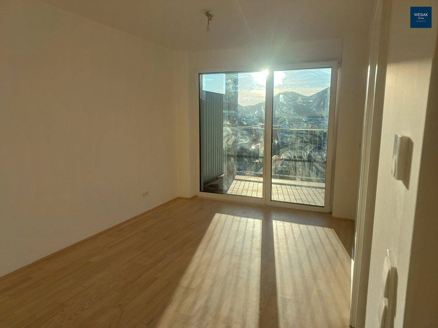 Lichtdurchflutetes Zimmer mit Holzboden und großer Schiebetür zum Balkon, ideal für viel Tageslicht.
