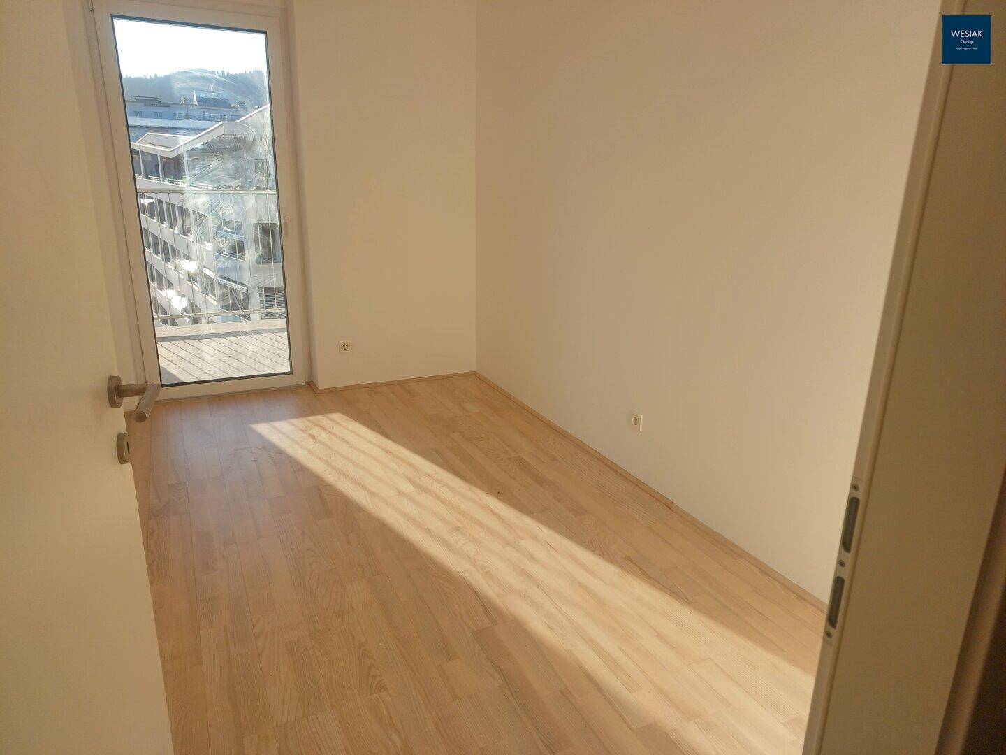 Freundliches Zimmer mit hellem Holzboden und Zugang zu einem Außenbereich, gut beleuchtet.