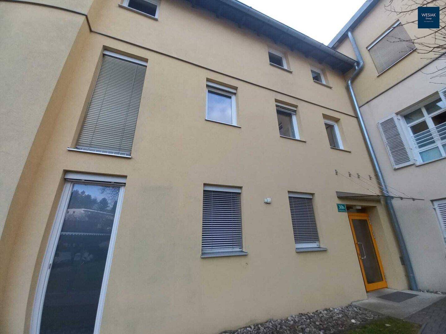 Nahaufnahme der gelben Gebäudefassade mit Fenstern, Jalousien und einem Eingangsbereich.