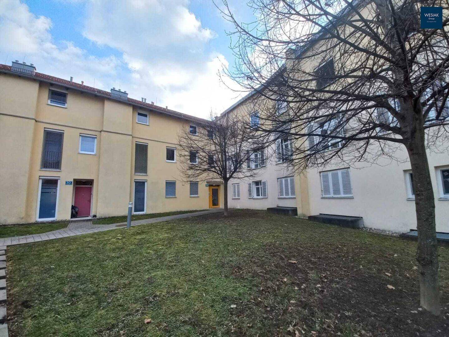 Wohngebäude mit gelber Fassade und einem Innenhof mit Bäumen und Rasenfläche unter bewölktem Himmel.