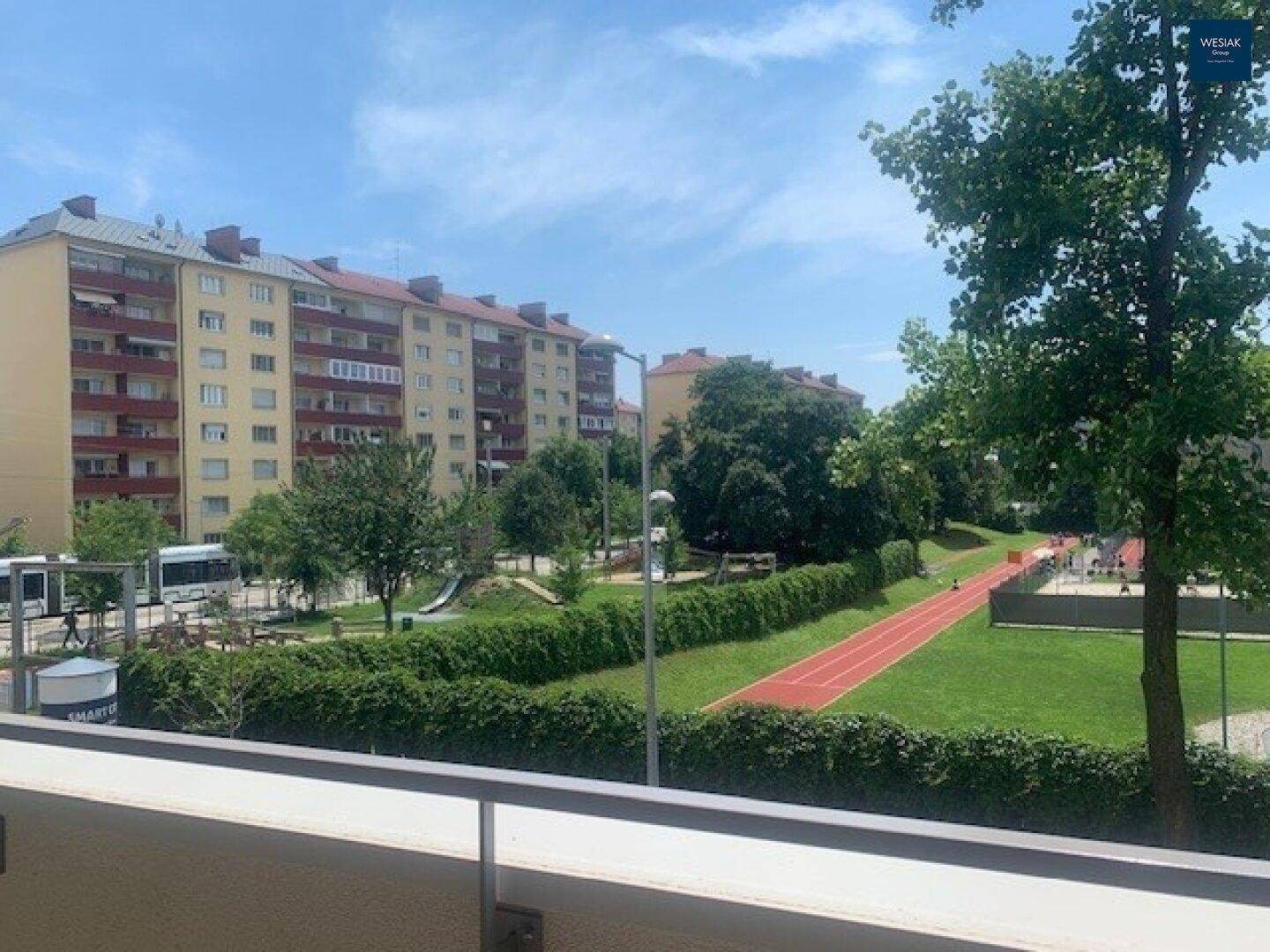 Blick vom Balkon auf eine grüne Parkanlage mit Spielplatz und Laufbahn, umgeben von Wohngebäuden.