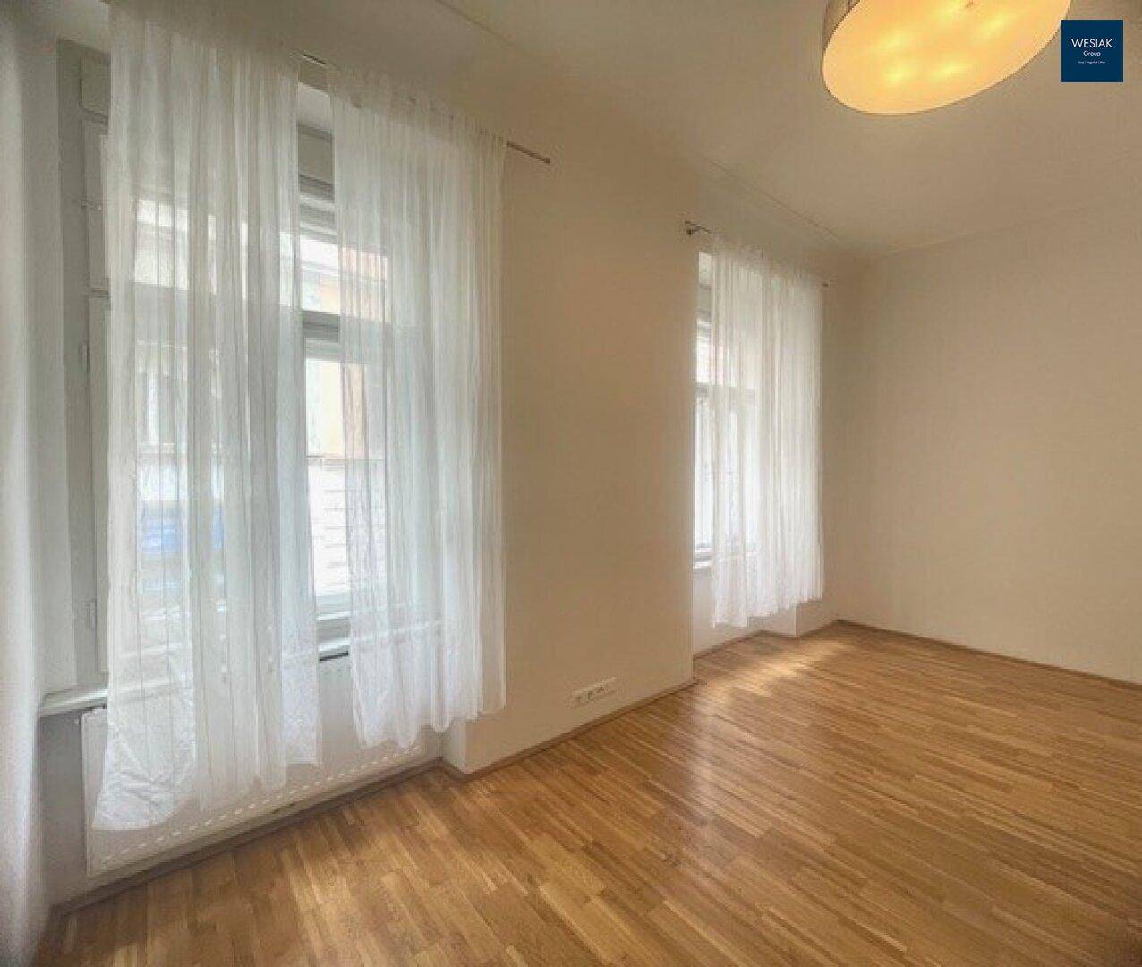Geräumiges Zimmer mit Parkettboden und zwei großen Fenstern, die viel Tageslicht hereinlassen.
