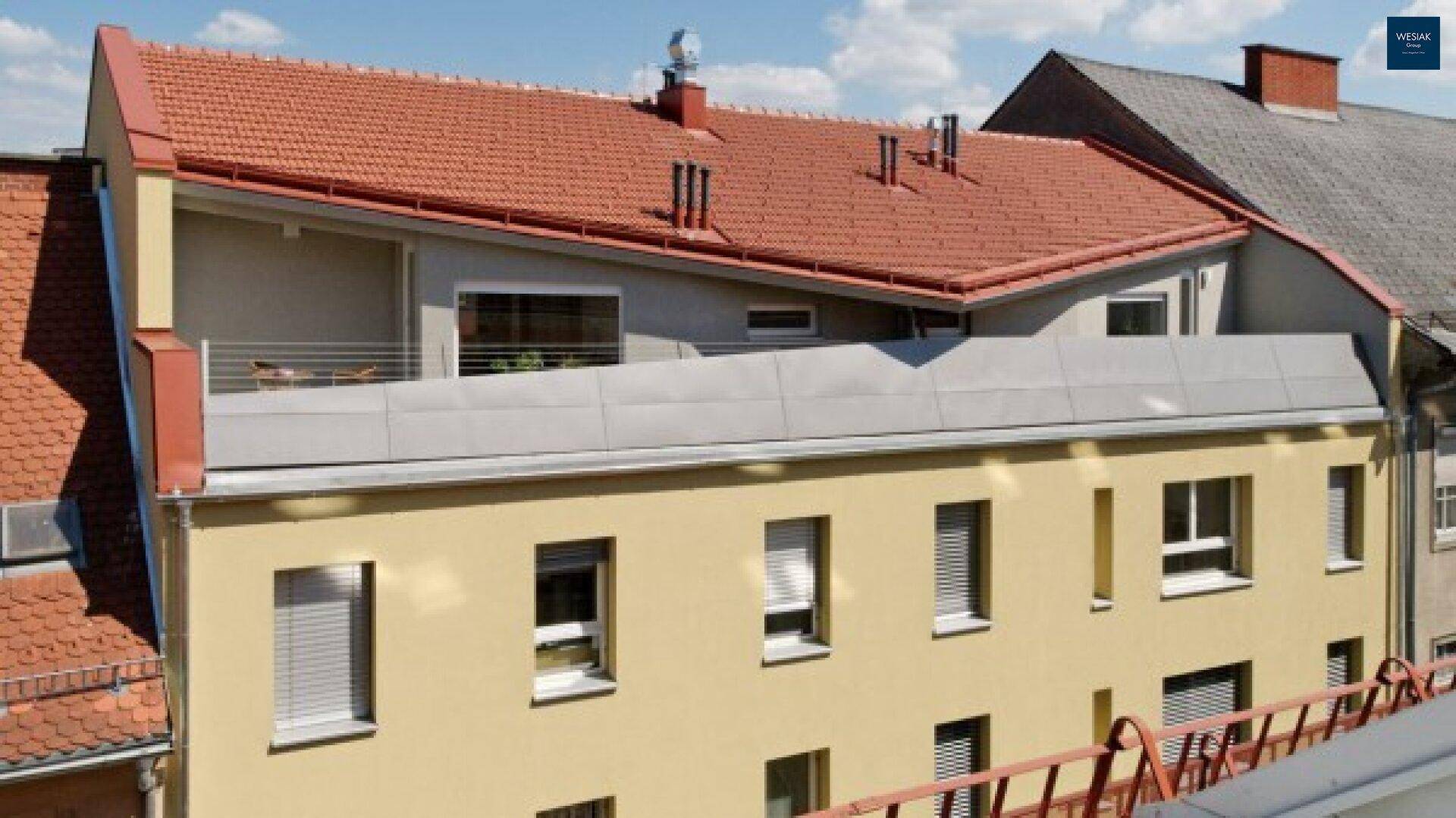 Ansicht eines Gebäudes mit rotem Ziegeldach und gelber Fassade, inklusive Balkonen im Obergeschoss.
