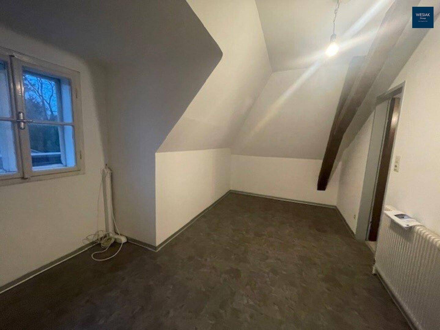 Helles Dachzimmer mit schrägen Wänden, sichtbarem Holzbalken und Fenster, ideal als Schlaf- oder Arbeitszimmer.