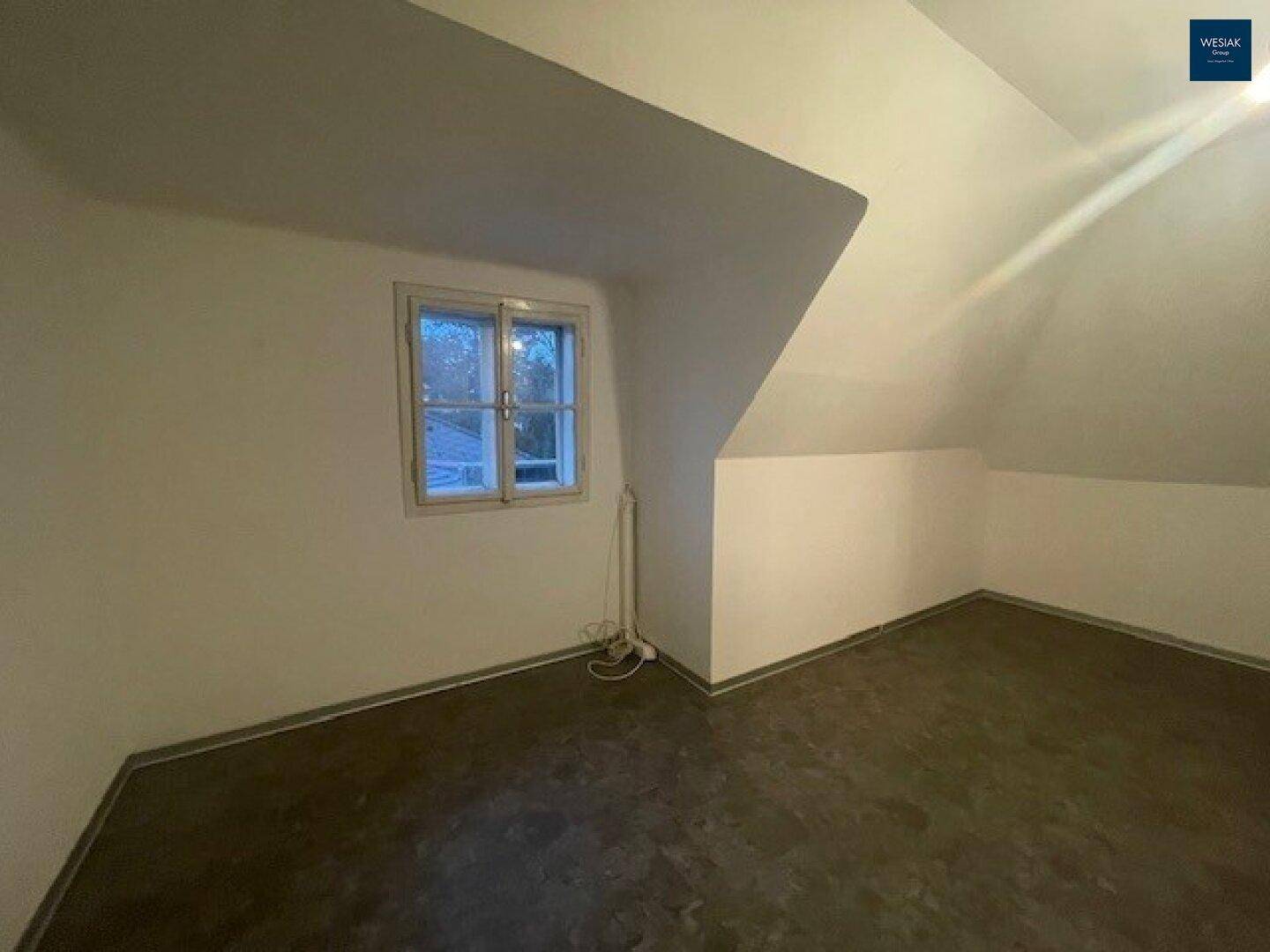 Geräumiges Dachzimmer mit schrägen Wänden und einem Fenster, das viel Tageslicht hereinlässt.