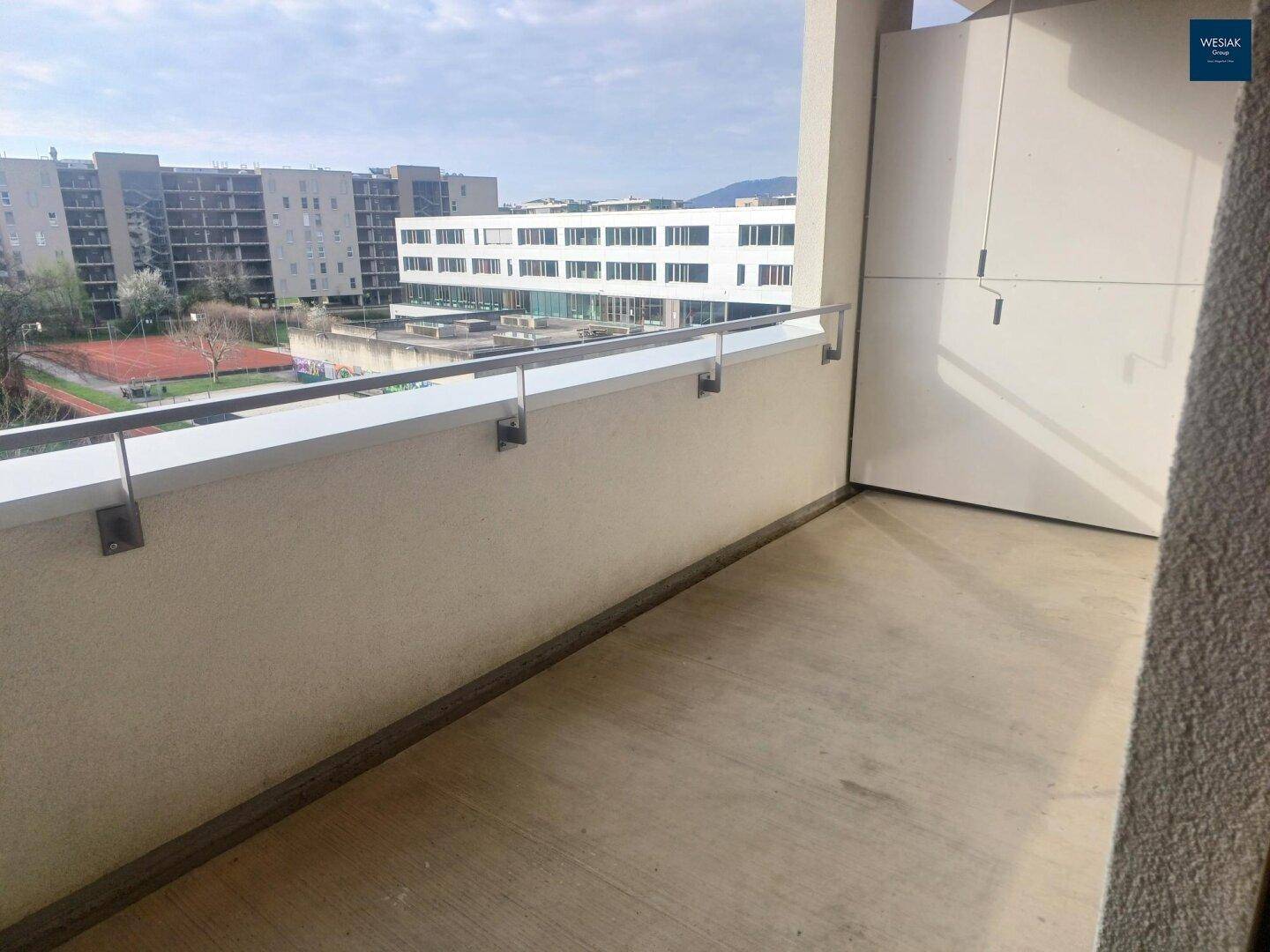 Heller Balkon mit Metallgeländer und Blick auf umliegende Gebäude und einen Tennisplatz.