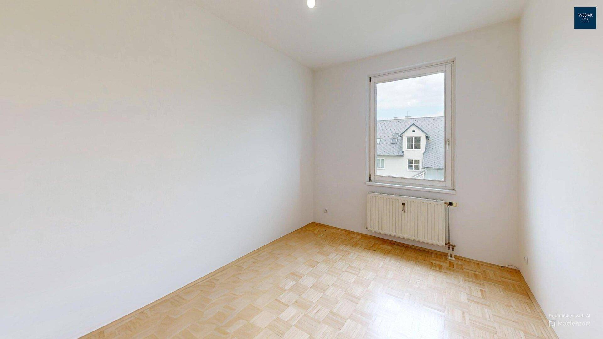 Leeres Schlafzimmer mit Parkettboden und Fenster mit Blick auf die Dächer.