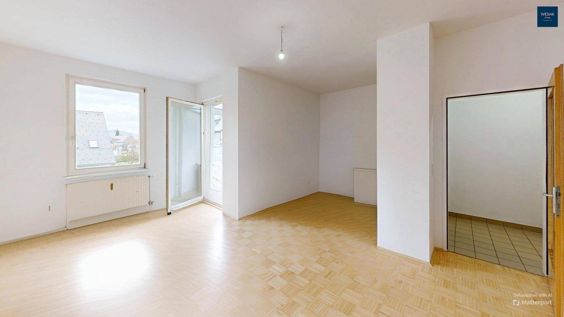 Leerer Wohnbereich mit Parkettboden und Zugang zum Balkon.