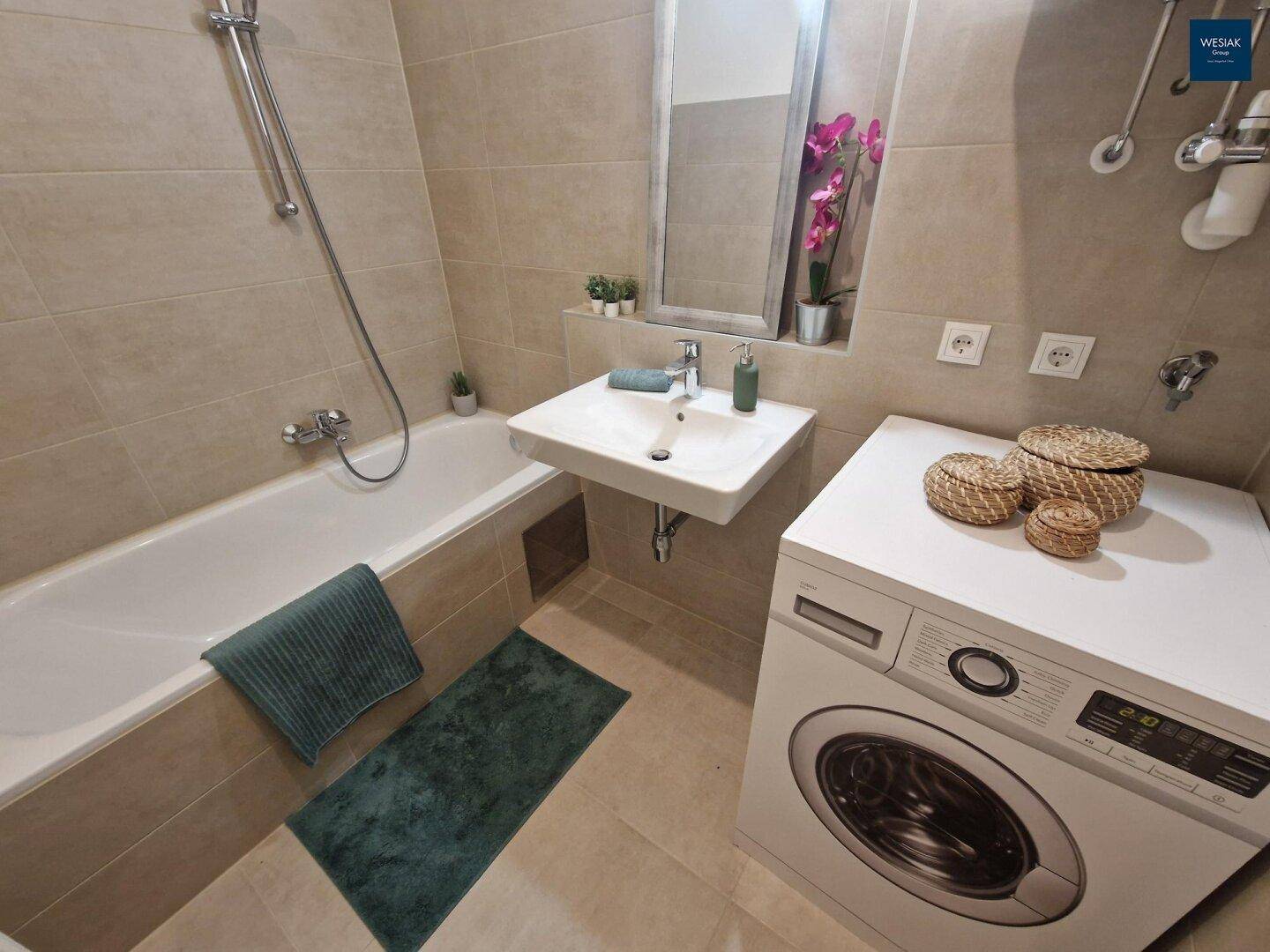 Badezimmer mit Badewanne, Waschbecken und Waschmaschine, gefliest in neutralen Tönen.