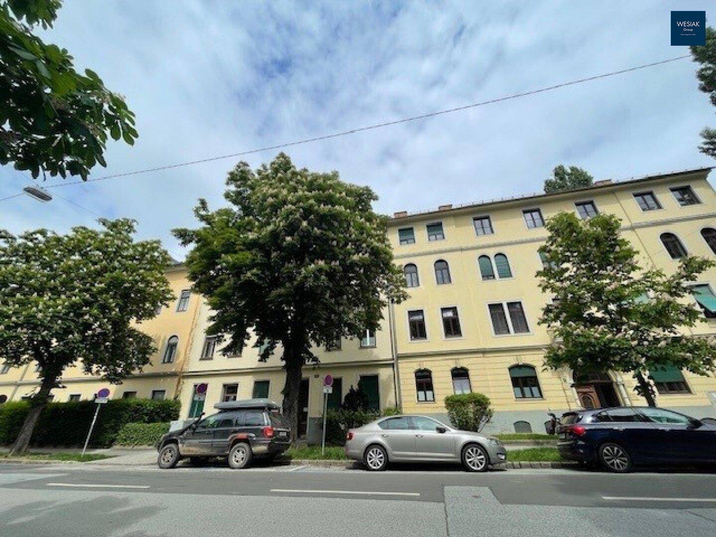 Ansicht eines älteren Wohngebäudes mit beiger Fassade, umgeben von üppigem Grün und parkenden Autos.