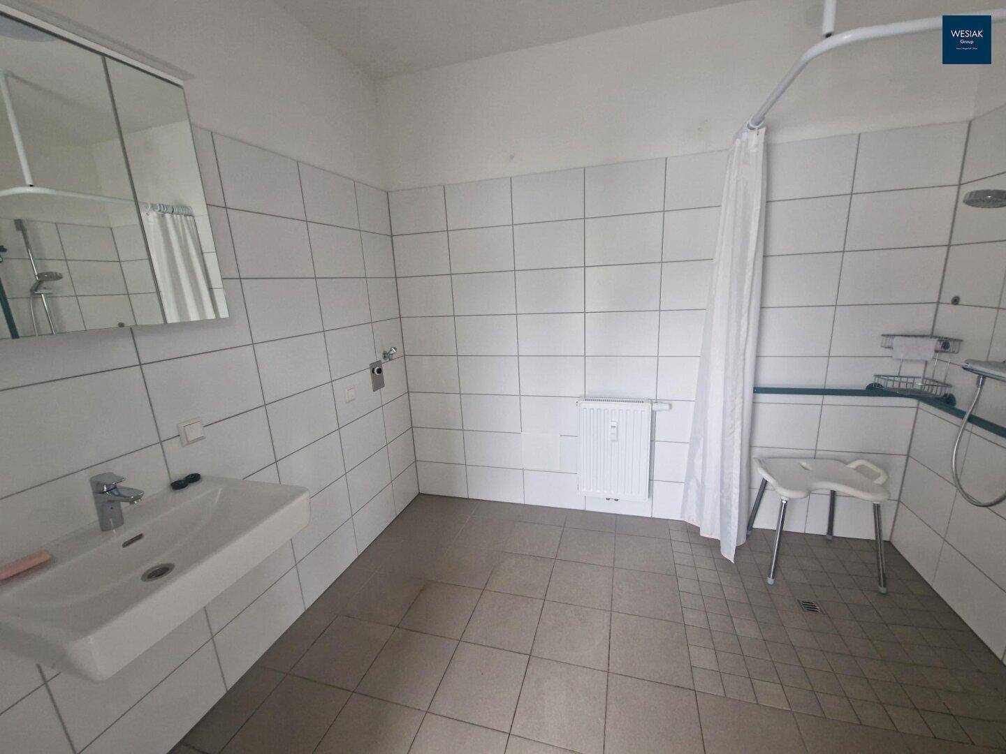 Barrierefreies Badezimmer mit Fliesenboden, Dusche und Haltegriffen.