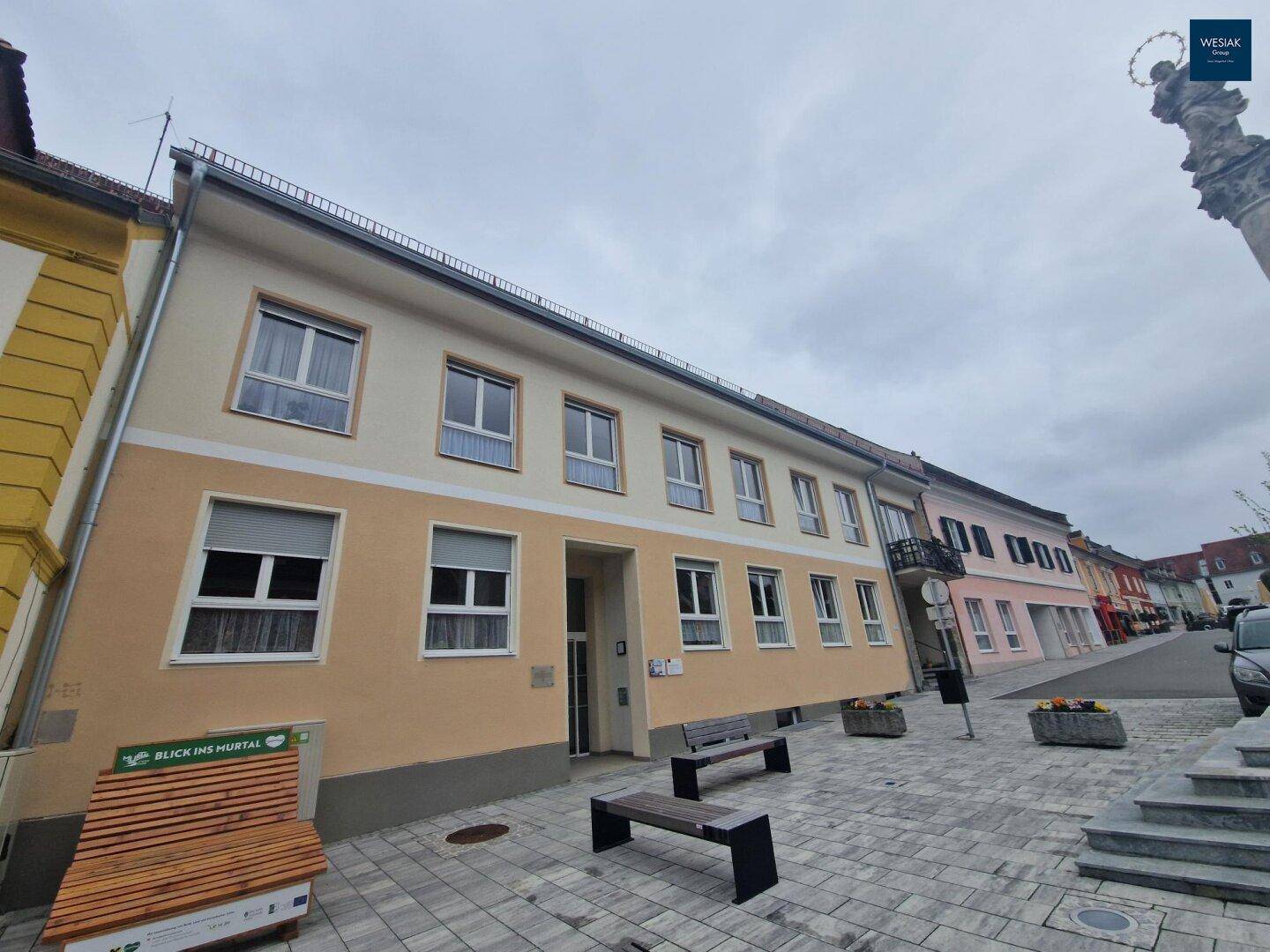 Fassade eines Gebäudes an einer gepflasterten Straße mit Bänken und einem Denkmal.