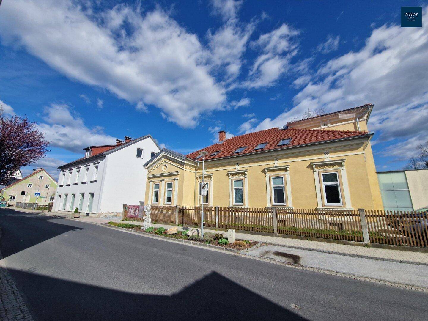 Gelbes Gebäude mit rotem Dach und Holzzaun an einer Straße unter blauem Himmel.