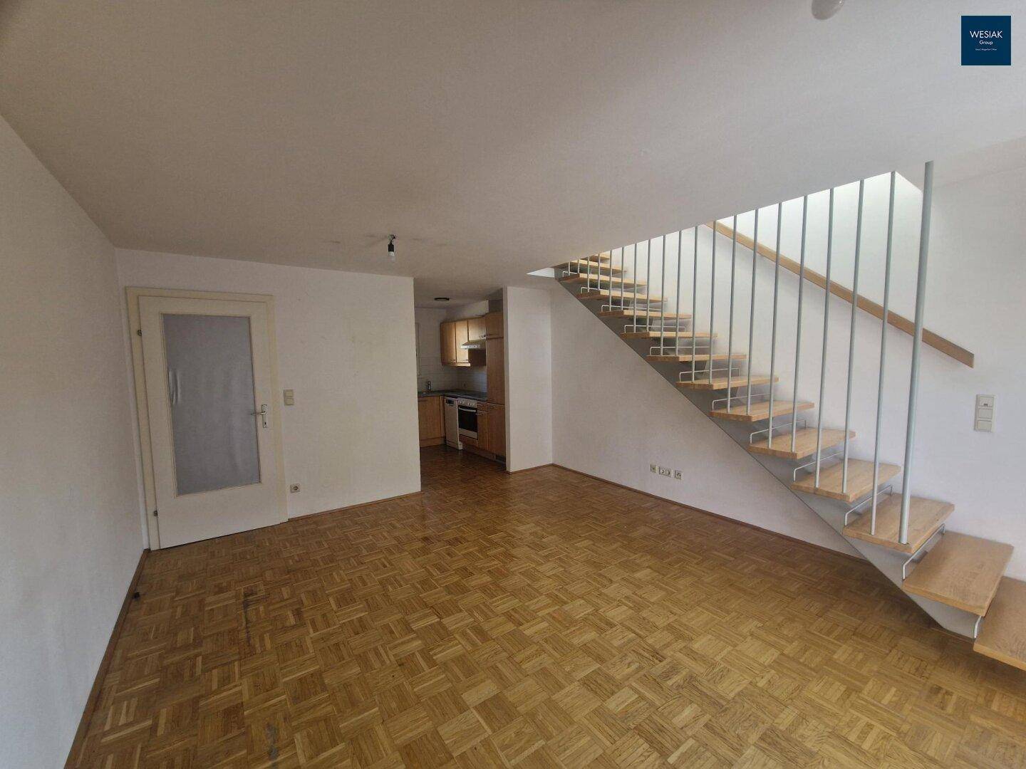 Offener Wohnbereich mit Parkettboden und Treppe, angrenzend an eine leere Küchennische.