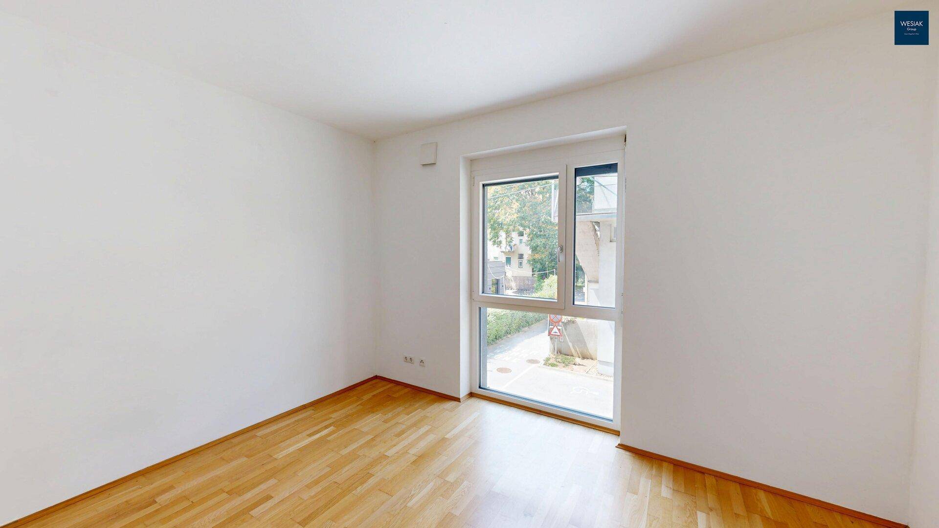 Leeres Zimmer mit Holzboden und großem Fenster mit Blick ins Grüne.