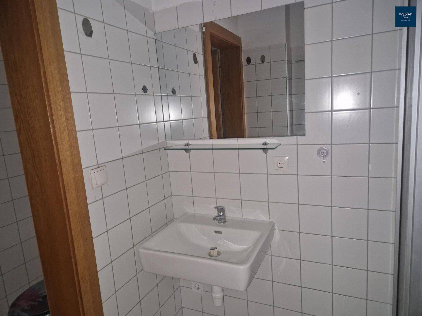 Badezimmer mit weißen Fliesen, Waschbecken, Spiegel und Holztürrahmen.