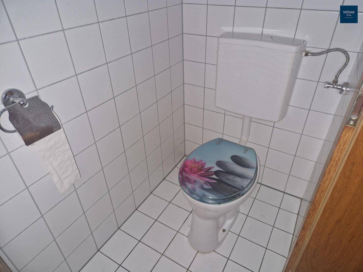 Separate Toilette mit weißen Fliesen und einem dekorativen Toilettendeckel.