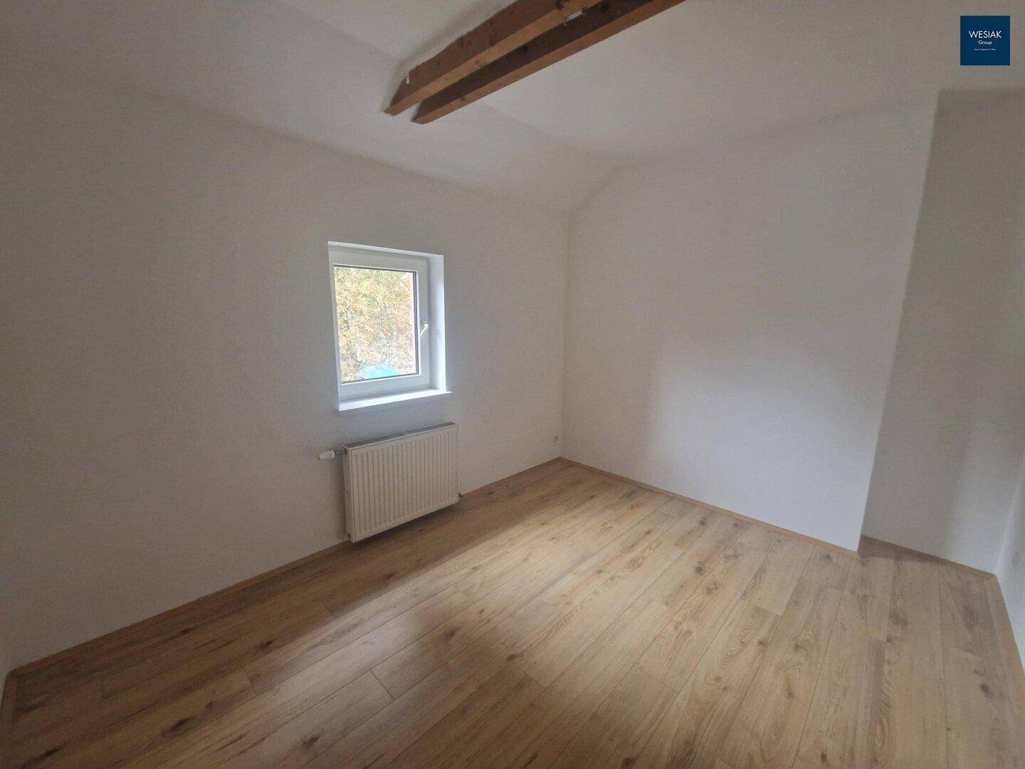 Leeres Zimmer mit Laminatboden, einem Fenster und sichtbaren Holzbalken an der Decke.