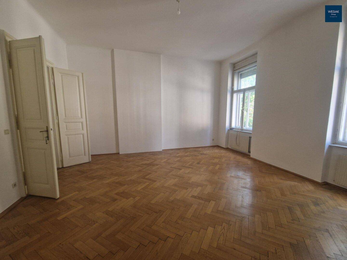 Großes Wohnzimmer mit Fischgrätparkett, hohen Fenstern und viel Tageslicht, ideal für eine helle Atmosphäre.