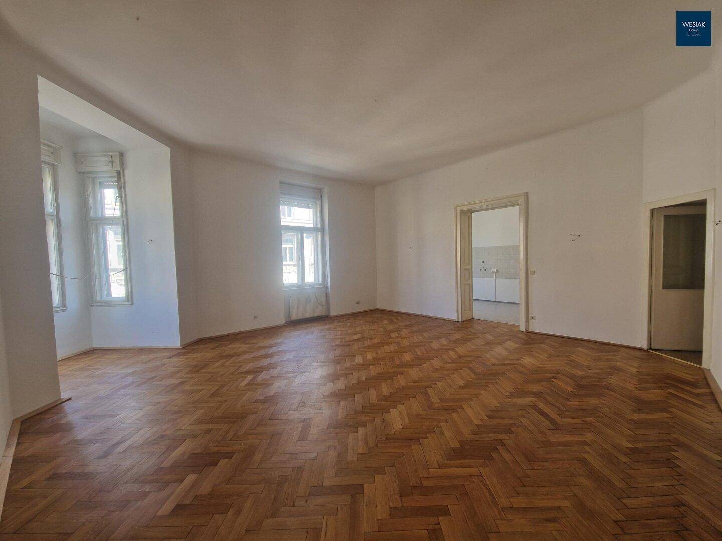 Großes Zimmer mit Fischgrätparkett, hohen Decken und Erkerfenstern, die viel Licht hereinlassen.