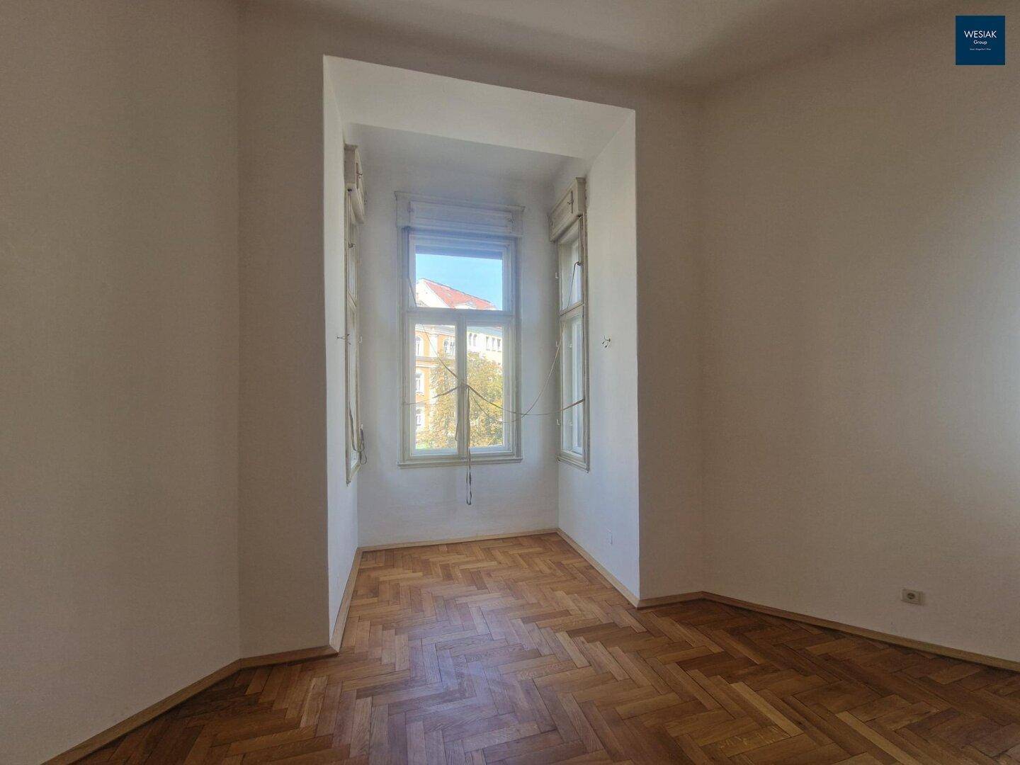 Kleines Zimmer mit Fischgrätparkett und einem großen Fenster, das viel Tageslicht spendet.