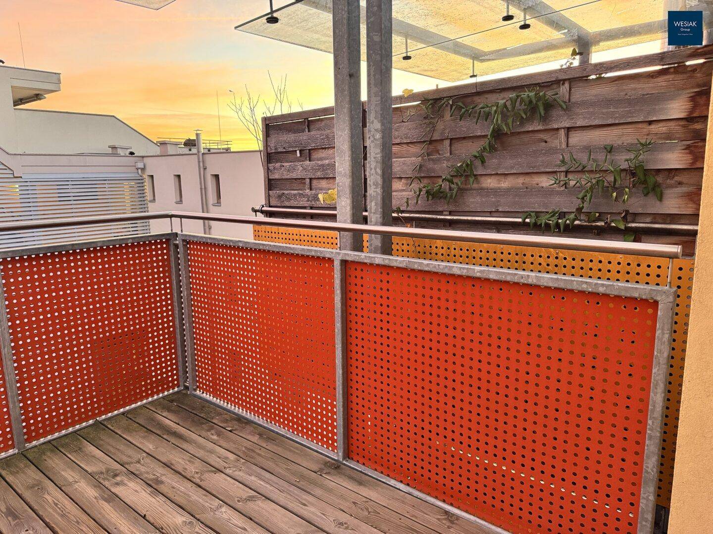 Balkon mit Holzboden und orangefarbenen Lochblech-Geländern bei Sonnenuntergang mit Blick auf die Umgebung.