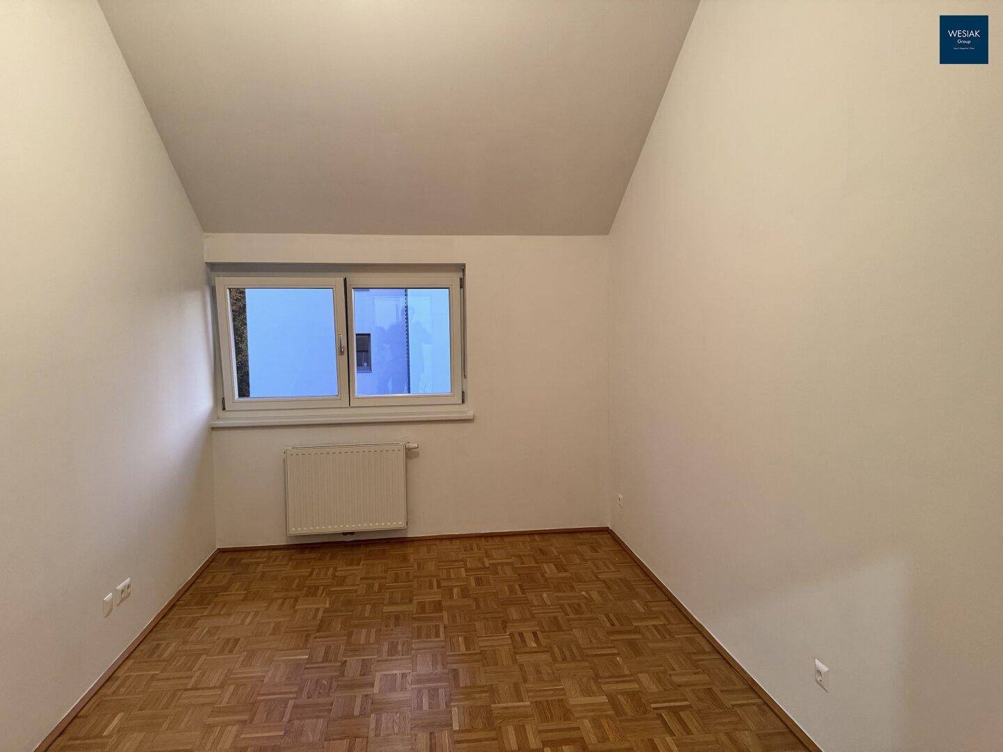 Helles Zimmer mit Parkettboden, Dachschräge und einem Fenster, das Tageslicht hereinlässt.