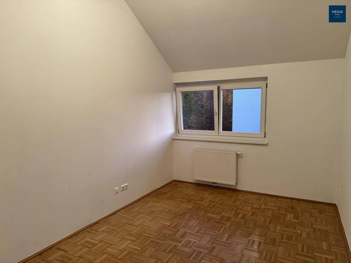 Geräumiges Zimmer mit Parkettboden, Dachschräge und einem Fenster mit Blick ins Freie.