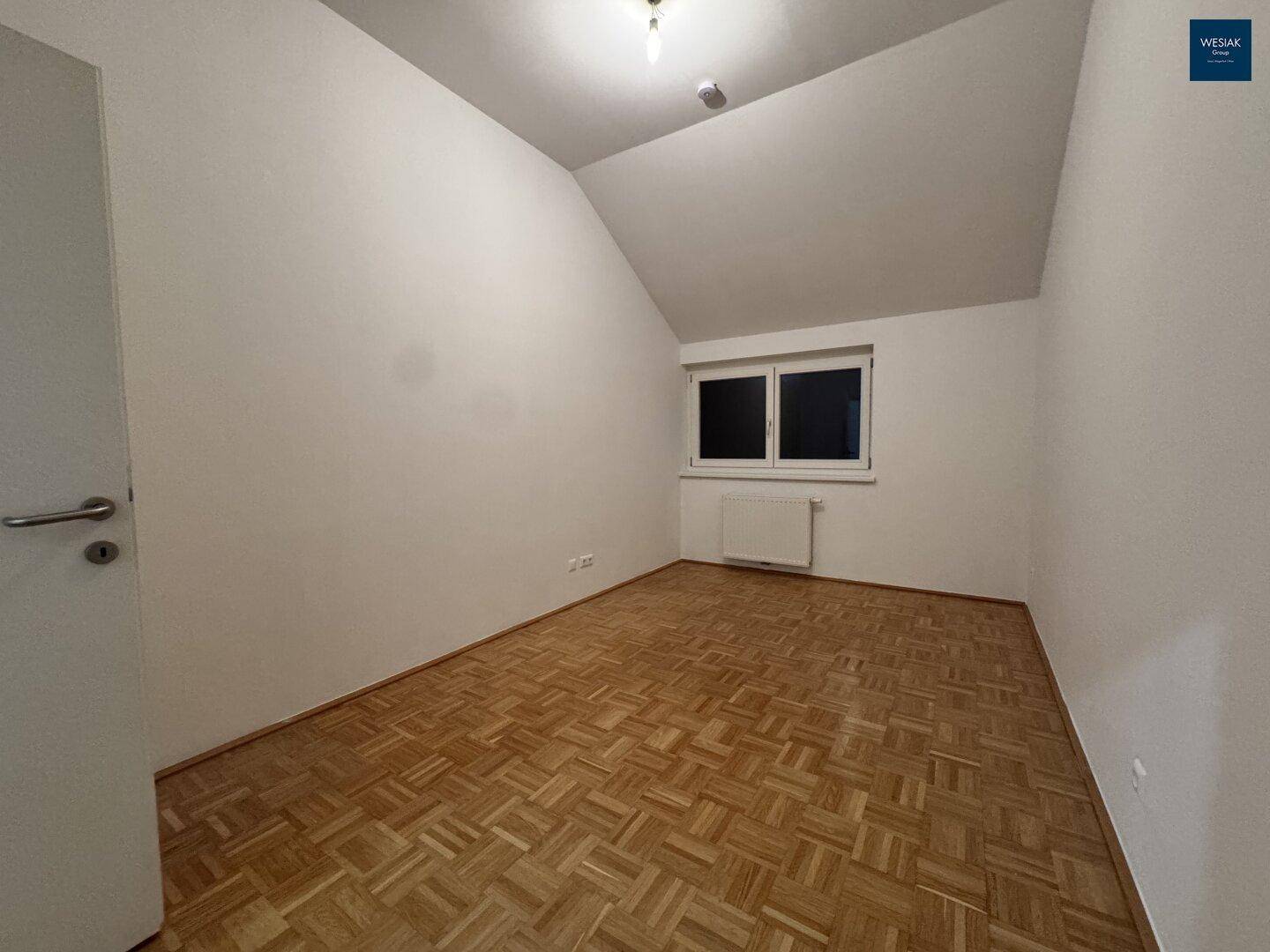 Leeres Zimmer mit Parkettboden, Dachschräge und einem Fenster, bereit zur individuellen Gestaltung.