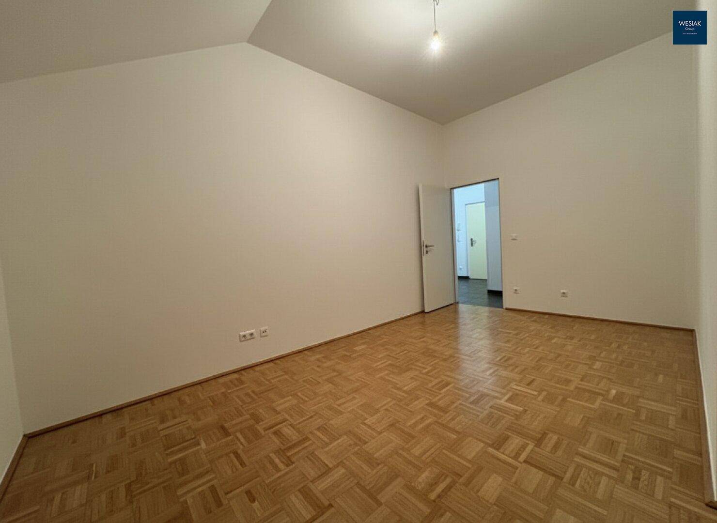 Helles Zimmer mit Parkettboden und einer offenen Tür, die den Blick in einen weiteren Raum freigibt.