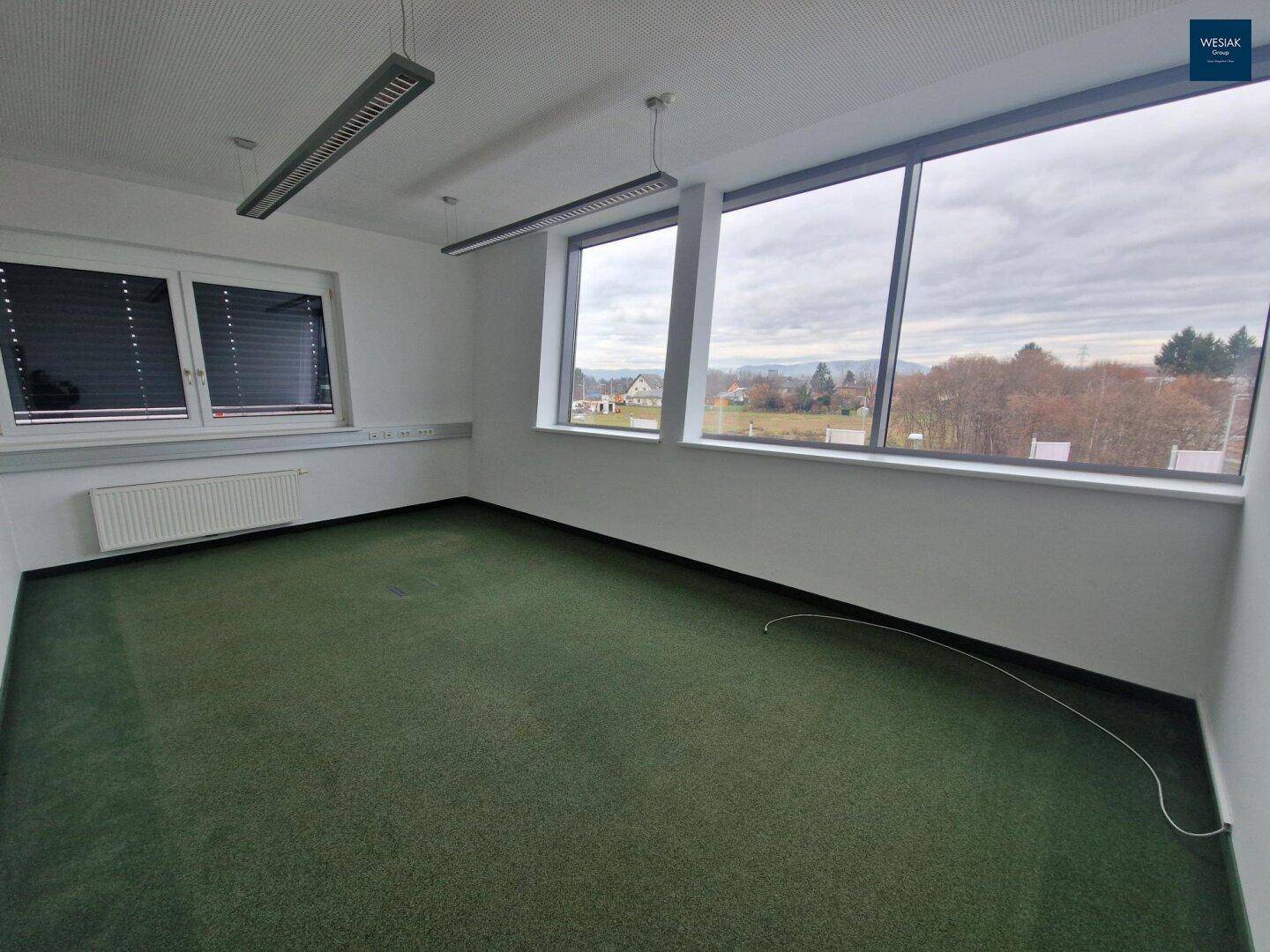 Leeres Büro mit Teppichboden, großen Fenstern und Blick auf die Landschaft und umliegende Gebäude.