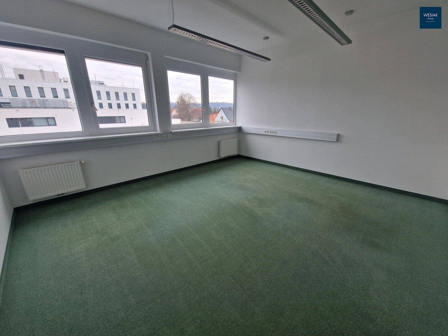 Leeres Büro mit Teppichboden, breiten Fenstern und Heizkörpern unter den Fensterbänken.