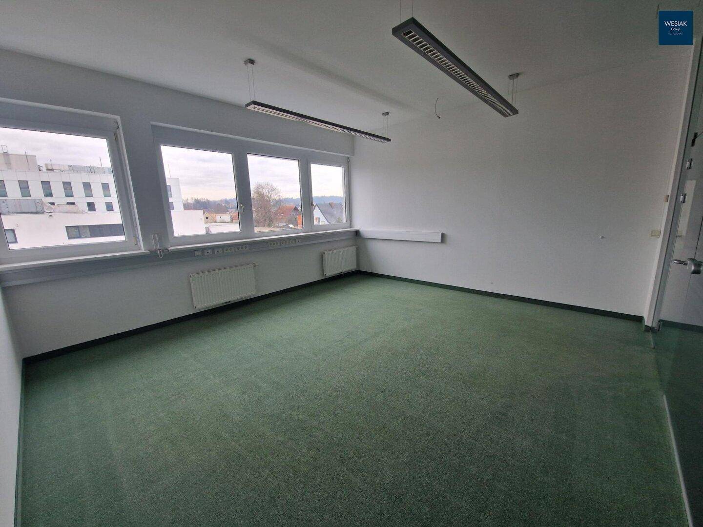 Geräumiges, leeres Büro mit Teppichboden, mehreren Fenstern und Blick auf die Stadt.