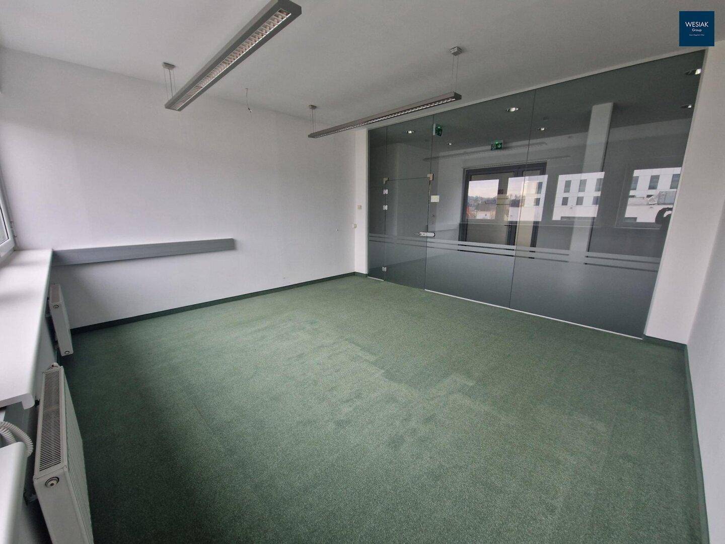 Leeres Büro mit Teppichboden, Glaswand und Heizkörper unter dem Fenster.